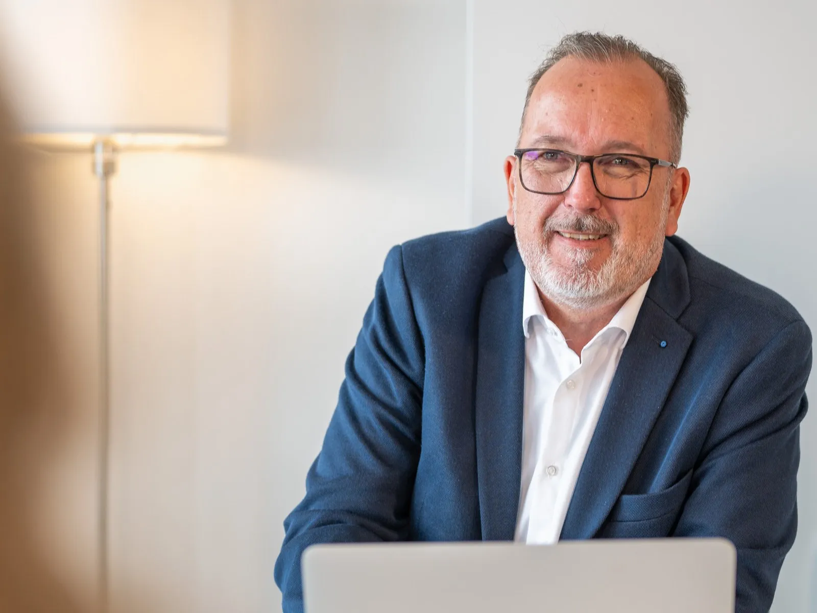 Björn-Olaf Lange sitzt lächelnd am Laptop und hört seinem Gegenüber zu, im Hintergrund eine warme Stehlampe